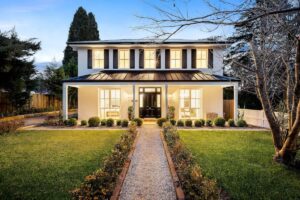 Two-storey weatherboard house lit at dusk, with a hedged path across the front lawn