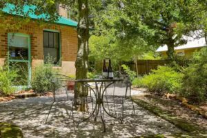 Wrought-iron garden table set with wine, under trees beside a brick cottage