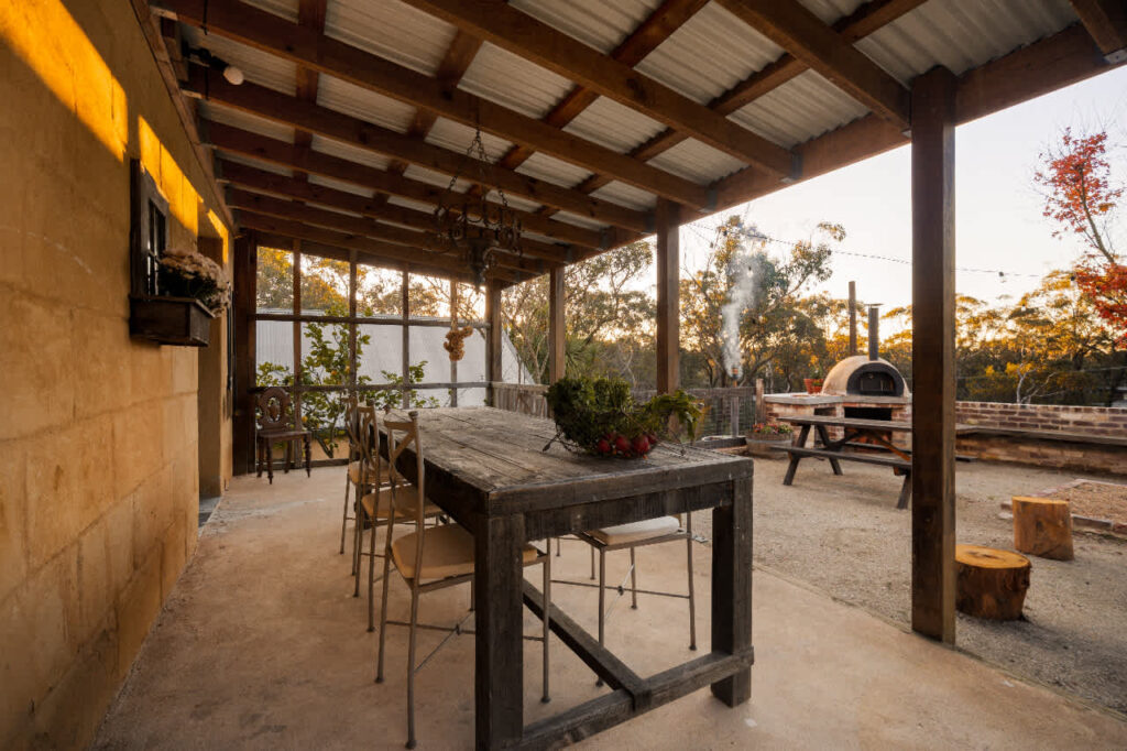 Covered outdoor dining area with a rustic table and wood-fired pizza oven at Harvest Cottage