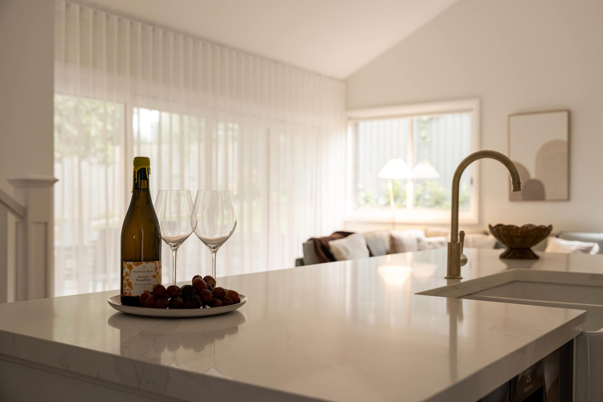Open-plan kitchen island set with a bottle of wine, two glasses and a bowl of cherries, looking through to a sunlit living area